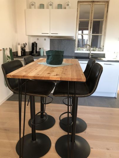 Een moderne eetruimte met een hoge houten tafel en vier zwarte barkrukken. Op de achtergrond zijn keukenkasten en een grote vensterpartij zichtbaar.