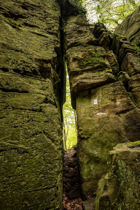 Ein schmaler Durchgang zwischen hohen Felsen im Wald. Die Steine sind grünlich und von Moos bedeckt.