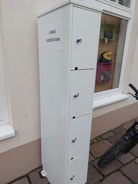 Eine E-Bike-Ladestation mit mehreren Fächern steht an der Wand. Neben der Station ist ein Fahrrad abgestellt.