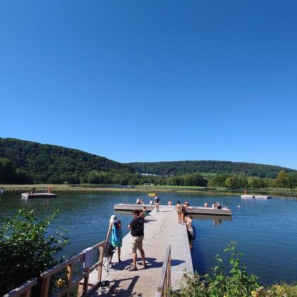 Un lac paisible sous un ciel bleu clair. Des gens profitent du soleil et de l'eau sur une jetée en bois.