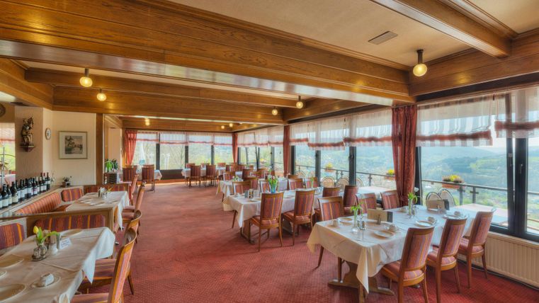 Ein elegantes Restaurant mit Holzelementen und großen Fenstern. Es sind gedeckte Tische und Stühle auf rotem Teppich zu sehen.