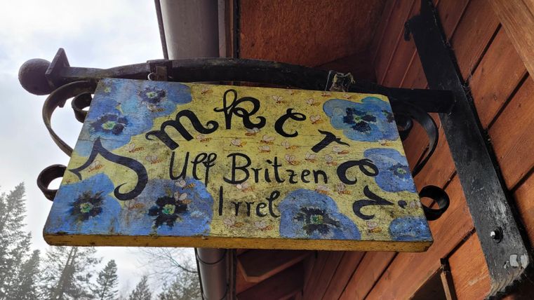 Ein handgemaltes Schild mit blauen Blumen und dem Text "Imkerei auf Brütten Irrel". Es hängt an einem Holzgebäude und vermittelt einen rustikalen Charme.