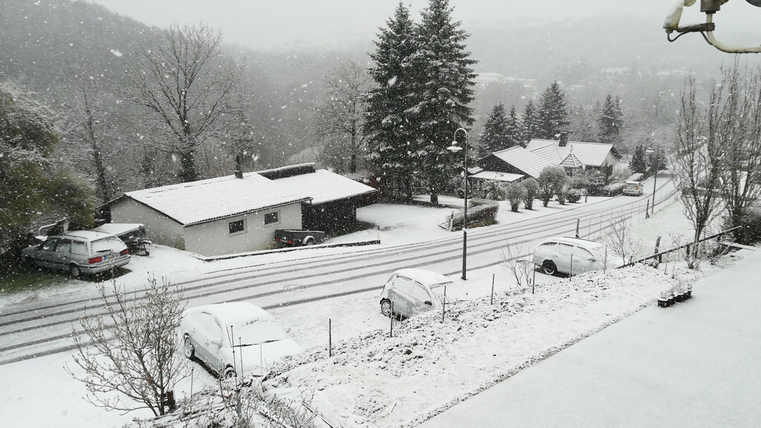 Eine verschneite Landschaft mit Häusern und parkenden Autos. Der Schnee fällt dicht und verdeckt die Straße.