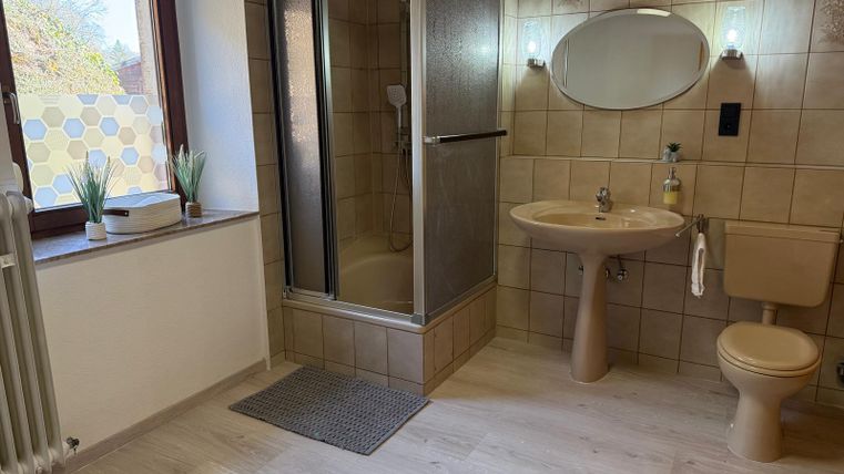 Een moderne badkamer met een douche, een wastafel en een toilet. De muren zijn betegeld en er is een raam met planten.
