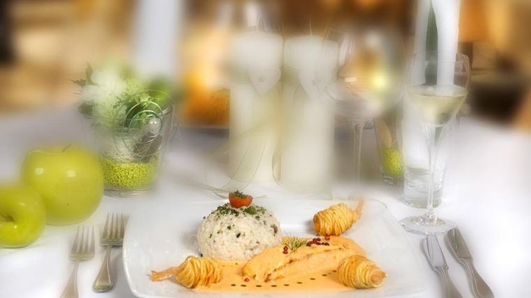 Ein elegantes Gericht mit Lachs, Reis und einer kleinen Tomate auf einem weißen Teller. Im Hintergrund sind Gläser und grüne Äpfel zu sehen.