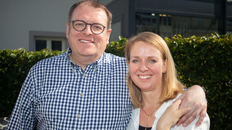 Ein lächelndes Paar steht in einem sonnigen Außenbereich. Im Hintergrund sind Pflanzen und ein modernes Gebäude zu sehen.