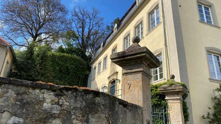 Ein historisches Gebäude mit hellen Wänden und großen Fenstern. Im Vordergrund ist eine alte Mauer mit einem Tor zu sehen.
