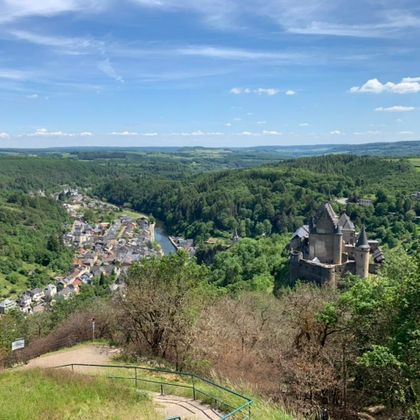 Un paysage pittoresque avec une rivière entourée de forêts vertes et de collines. Au premier plan se trouve un vieux château qui domine la scène.