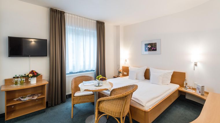 Ein gemütliches Hotelzimmer mit einem Doppelbett, einem Tisch und zwei Stühlen. Es gibt einen Fernseher und ein Fenster mit Vorhängen, die viel Licht hereinlassen.