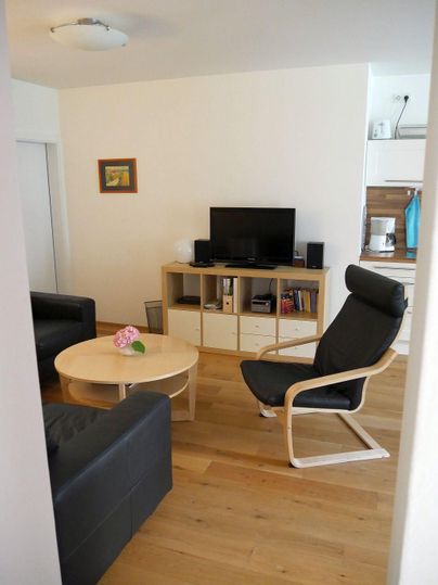 Ein modernes Wohnzimmer mit einem schwarzen Sofa und einem komfortablen Stuhl. Der Raum ist hell und einladend, mit einem Tisch und einem Fernseher.