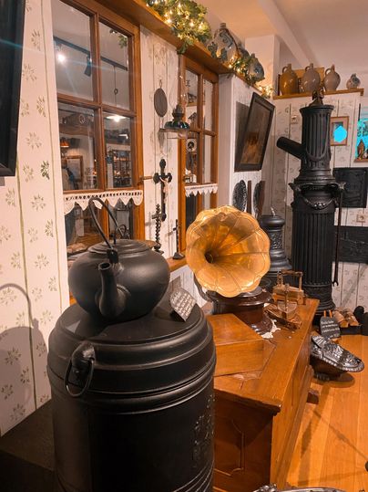 Ein traditionelles Zimmer mit Antiquitäten, darunter ein schwarzer Kessel und ein Grammophon. Die Wände sind mit floralen Mustern dekoriert und es gibt Holzgegenstände im Raum.