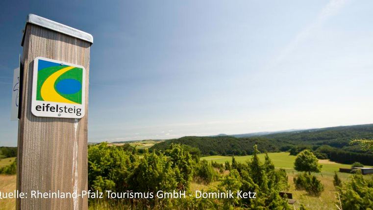 Een bewegwijzering van de Eifelsteig staat voor een uitgestrekt, groen landschap. De lucht is helder en blauw.