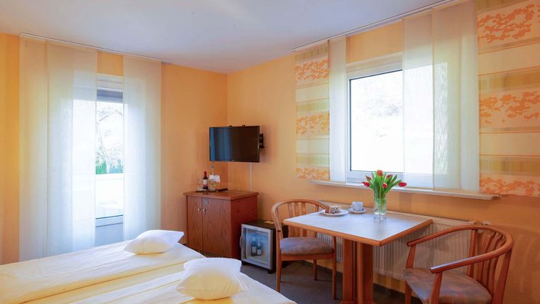 Ein gemütliches Hotelzimmer mit zwei Einzelbetten, einem Fernseher und einem kleinen Tisch. Große Fenster lassen viel Licht herein und die Wände sind in warmen Farben gestrichen.