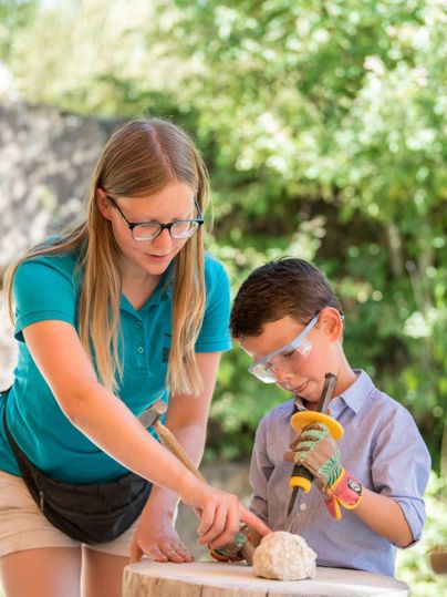 Ein Junge und ein Mädchen arbeiten gemeinsam an einem Holzblock. Beide tragen Schutzbrillen und konzentrieren sich auf ihre Aufgabe.
