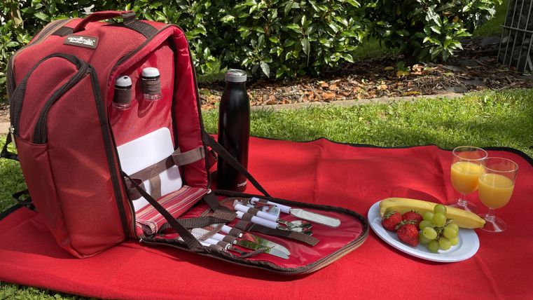 Un sac à dos de pique-nique rouge est posé sur une couverture dans la verdure. Il contient des couverts, des bouteilles et un espace avec des fruits frais et des boissons.