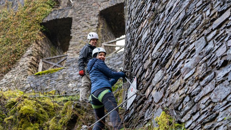 Two people climbing on an old stone wall. The surroundings are green and wooded, with a historic building in the background.