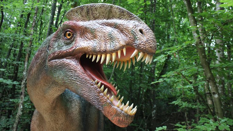 Ein prähistorisches Dinosauriermodell mit geöffnetem Maul steht in einem dichten Wald. Die Umgebung ist grün und üppig.