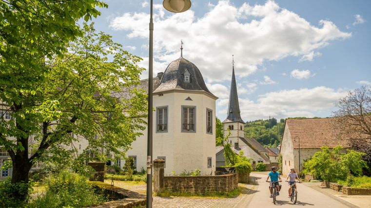 Zwei Radfahrer auf einer Straße in Wolsfeld, umgeben von historischen Gebäuden und einer Kirche im Hintergrund.