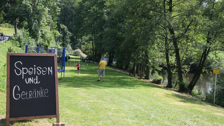 Ein kleiner Park am Fluss mit einer Wiese und schattigen Bäumen. Ein Schild zeigt "Speisen und Getränke" an.