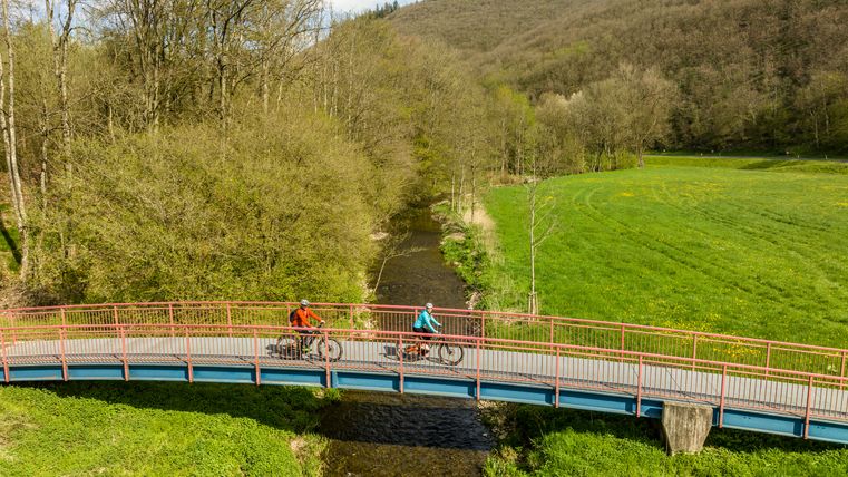 Zwei Radfahrer auf einer Brücke über einen Fluss, umgeben von grünen Wiesen und bewaldeten Hügeln.