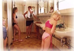 Eine helle Umkleidekabine mit mehreren Personen in Badebekleidung. Zwei Frauen unterhalten sich, während ein Mädchen am Waschbecken steht.