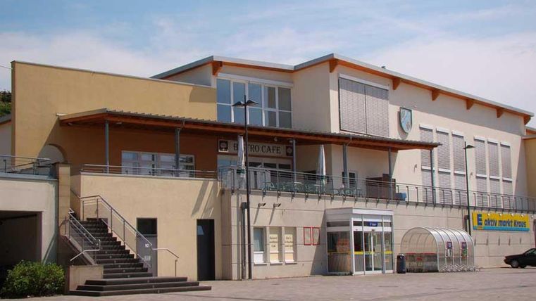 Ein modernes Gebäude mit einer großen Terrasse und einem Eingangsbereich. Vor dem Gebäude befinden sich Stufen und eine gepflasterte Fläche.