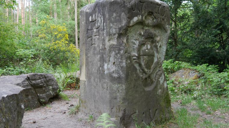 Ein großer, bearbeiteter Stein steht inmitten eines dichten Waldes. Auf dem Stein sind kunstvolle Reliefs zu sehen, umgeben von grüner Vegetation.