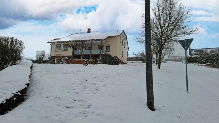 Ein Haus im Schnee mit einer schneebedeckten Landschaft. Der Himmel ist bewölkt und es gibt einige Bäume in der Umgebung.