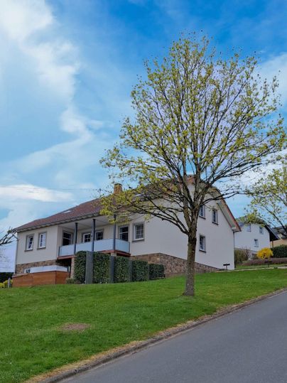 Ein modernes Haus auf einem Hang mit einem grünen Rasen und einem Baum. Der Himmel ist blau mit vereinzelten Wolken.