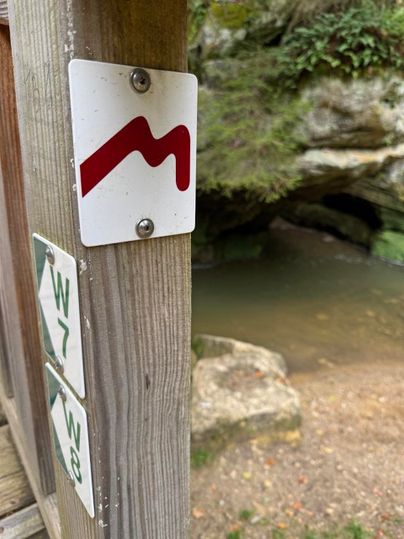 Een wegwijzer met een rood symbool op een houten paal. Op de achtergrond is een klein watergebied en rotsen te zien.