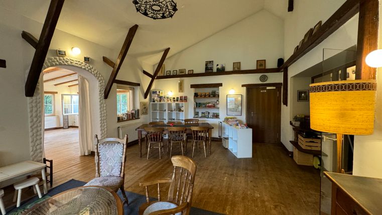 Un salon lumineux et accueillant avec un sol en bois et des poutres en bois au plafond. On aperçoit un coin salon confortable et divers objets de décoration aux murs.