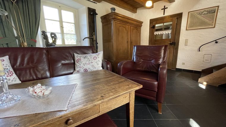 Ein gemütliches Wohnzimmer mit einem Holztisch, einem roten Ledersofa und einem Sessel. Helle Fenster und ein Holzregal schaffen ein einladendes Ambiente.