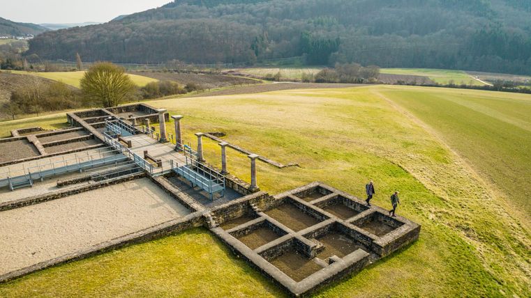 Eine archäologische Ruine auf einer grünen Wiese, umgeben von Hügeln. Zwei Personen erkunden die Überreste eines Gebäudes.