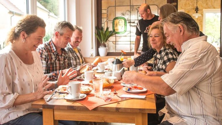Eine Gruppe von fünf Personen sitzt gemeinsam an einem Tisch und genießt ein Frühstück. Auf dem Tisch stehen Tassen, Teller mit Essen und frische Snacks.