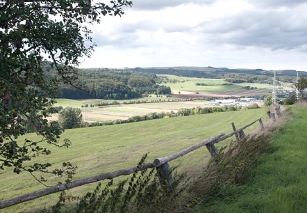 Landscape with meadows, trees and a wooden fence in the foreground. Fields and hills can be seen in the background under a cloudy sky., &copy; V. Teuschler