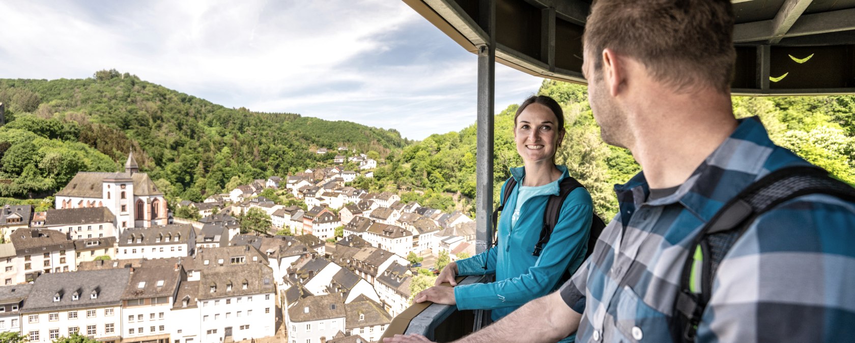 Zwei Personen stehen auf einem Aussichtspunkt und blicken auf eine malerische Stadt mit Kirche und gr&uuml;nen H&uuml;geln im Hintergrund., &copy; Eifel Tourismus GmbH, Dominik Ketz