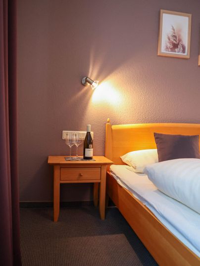 Ein gemütliches Schlafzimmer mit einem Holzbett und einem Nachttisch. Auf dem Nachttisch stehen zwei Gläser und eine Flasche Wein.