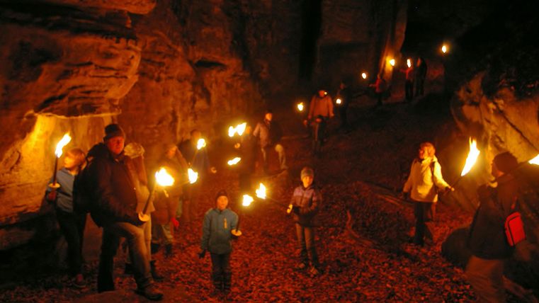 Eine Gruppe von Menschen geht mit Fackeln durch eine dunkle Höhle. Der Boden ist mit Laub bedeckt, und das Licht der Fackeln beleuchtet die Umgebung.