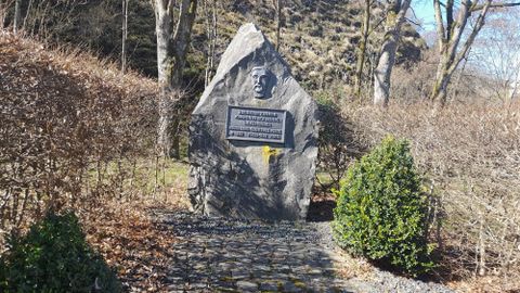 Een gedenksteen in een rustige omgeving, omgeven door struiken en kleine bomen. Op de steen staat een reliëf en een inscriptie.