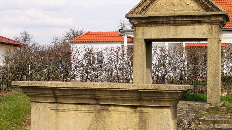 Nachbildung eines gallo-römischen Weihdenkmals mit Inschrift, umgeben von modernen Gebäuden und Büschen.