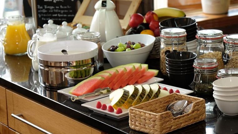 Ein schön arrangiertes Frühstücksbuffet mit frischem Obst, darunter Wassermelonen, Melonen und Trauben. Im Hintergrund stehen verschiedene Gläser und Behälter für weitere Leckereien.