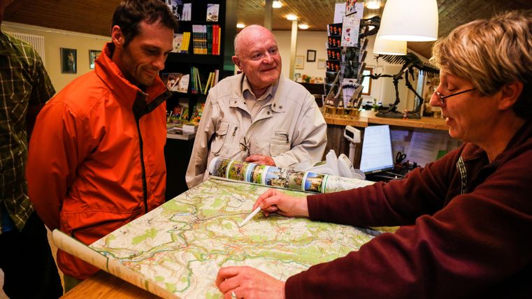 Ein Mann in einem orangefarbenen Jackett und ein älterer Herr hören einer Frau zu, die auf eine Karte zeigt. Die Szene spielt sich in einem gemütlichen Raum ab, der mit verschiedenen Gegenständen dekoriert ist.