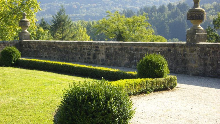 Ein gepflegter Garten mit rund geformten Büschen und einer Steinmauer. Im Hintergrund sind sanfte Hügel und üppige Bäume zu sehen.