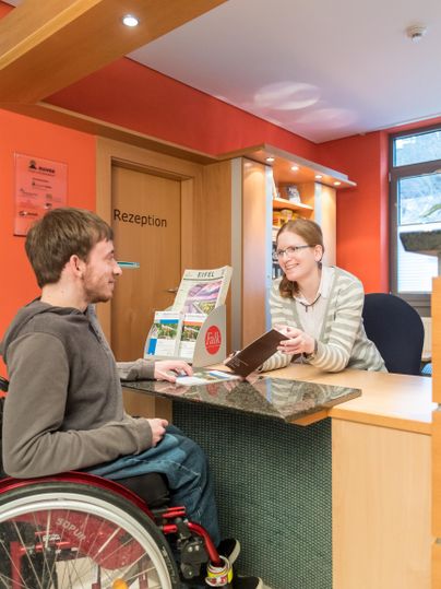 Ein Empfangsbereich mit einem Mann im Rollstuhl und einer Frau, die am Schalter steht. Beide scheinen ein freundliches Gespräch zu führen.