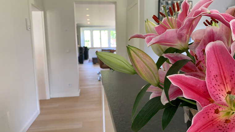 Ein heller Innenraum mit Holzfußboden und großen Fenstern. Im Vordergrund steht ein Blumengefäß mit rosa Lilien.