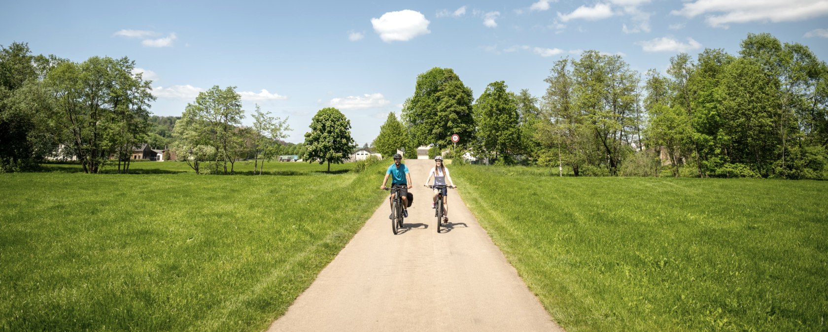 Deux cyclistes roulent sur une route asphaltée à travers un paysage verdoyant. Le ciel est bleu avec quelques nuages., © Eifel Tourismus GmbH, Dominik Ketz
