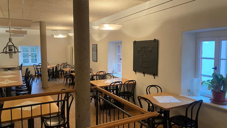 Ein gemütliches Restaurant mit Holzstühlen und Tischen. Die Wände sind hell gestrichen und es gibt große Fenster, die für viel Licht sorgen.