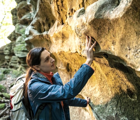 A woman in a blue anorak touches a sandstone wall in the forest. She is carrying a rucksack and looking at the structure of the stone., &copy; Eifel Tourismus GmbH, Dominik Ketz