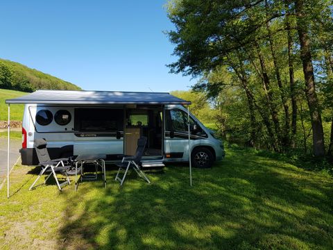 Ein Wohnmobil steht auf einer Wiese, umgeben von Bäumen. Zwei Klappstühle sind vor dem Fahrzeug aufgestellt, ideal zum Entspannen in der Natur.