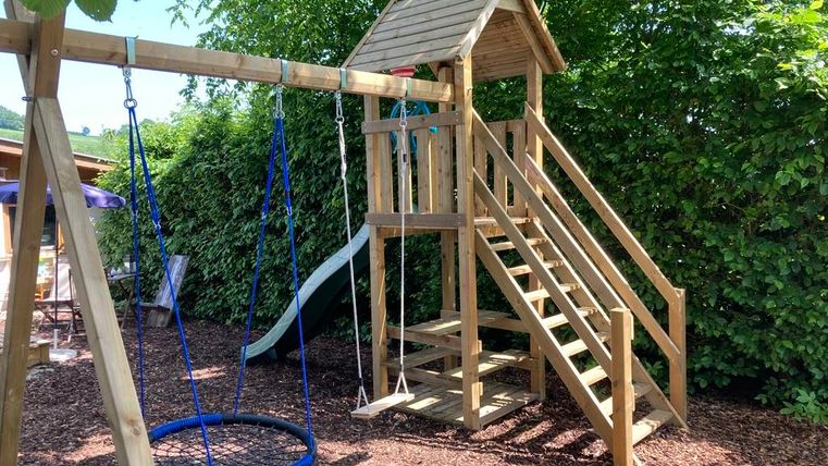 Een speeltuin met een houten glijbaan, een zandafgraving en schommels. De omgeving is omringd door groene planten.
